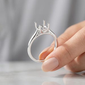 以下が含まれることがあります： 単一の宝石を保持するように設計された4本爪のヘッドを備えた、光沢のあるシルバー調のリングセッティング。バンドは滑らかで丸みを帯びており、爪を支える構造化されたギャラリーへと続いており、指輪のクラシックな土台を形成しています。