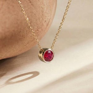 May include: A gold necklace with a round, faceted ruby pendant. The ruby is set in a gold bezel, and the chain is delicate. The necklace is displayed on a neutral background, highlighting the gemstone's vibrant red color.