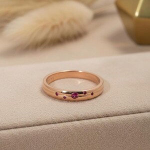 May include: A rose gold band ring featuring a polished finish and a horizontal groove detail. The ring is set with five small, round, vibrant pink gemstones of varying sizes arranged in a cluster along the front surface of the metal band.