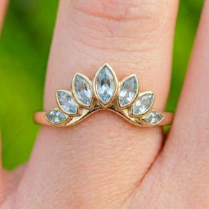 May include: A gold ring featuring a curved, crown-like band set with seven light blue, marquise-cut gemstones arranged in a fan shape. The metal band has a polished finish, and the stones are secured in individual prong settings along the top.