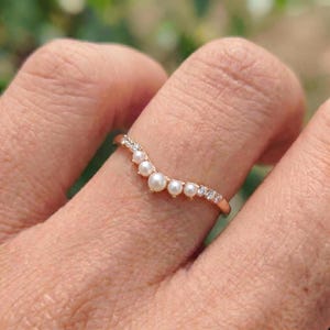 May include: A rose gold ring with a curved design. The ring features a row of small, round pearls and sparkling clear gemstones. The ring is on a finger.