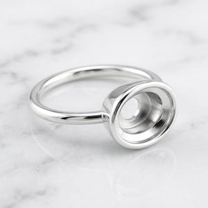 Puede incluir: Un anillo de tono plateado pulido con un engaste circular prominente en forma de copa, diseñado para sostener una piedra o cabujón. La banda metálica es lisa y redondeada, conectada a una montura cóncava y profunda.