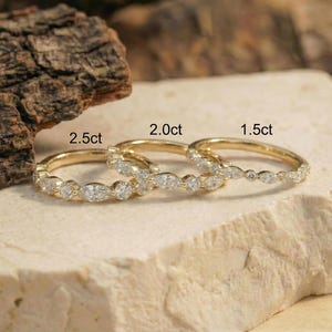 May include: Three gold rings with diamond accents are displayed. The rings are labeled 2.5ct, 2.0ct, and 1.5ct. The rings are set on a light-colored, textured surface, with a piece of wood in the background.