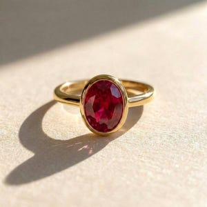 May include: A gold ring featuring a central oval-cut deep red gemstone set in a smooth bezel setting. The band is polished gold, creating a clean and simple design that highlights the vibrant color and faceted surface of the prominent red stone.