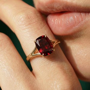 May include: A gold ring featuring a large, rectangular, deep red gemstone. The ring's band has a split design, and the gemstone is held in place by four prongs. The ring is shown on a finger.