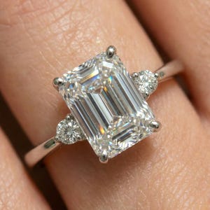 May include: A close-up of a diamond ring featuring a large, emerald-cut center stone flanked by two smaller round diamonds. The ring has a thin, silver band and is set against a blurred background.