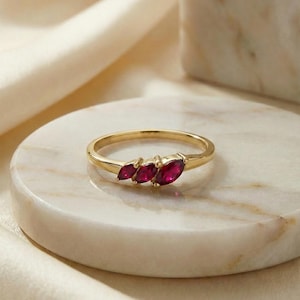 May include: A gold ring featuring three marquise-cut deep red gemstones arranged horizontally in a row. The band is thin and polished, showcasing a simple metal design that highlights the vibrant color and elongated shape of the three stones.