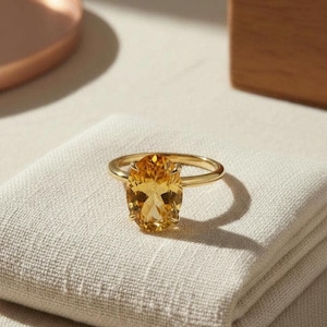 Könnte beinhalten: Ein goldener Ring mit einem großen, oval geschliffenen, gelben Edelstein. Der Ring liegt auf einem gefalteten, cremefarbenen Tuch. Der Edelstein ist der Blickfang, seine Facetten reflektieren das Licht.