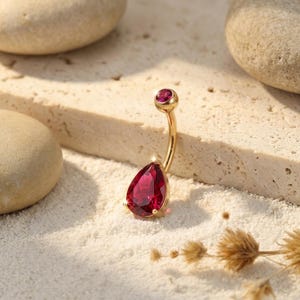 May include: A gold-toned curved barbell belly button ring featuring a small round red gemstone at the top and a larger pear-shaped red gemstone at the bottom. The jewelry has a polished metallic finish and is positioned upright on a textured surface.