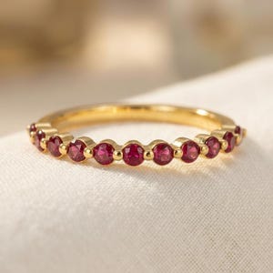 Può includere: Un anello a fascia in oro con una fila di piccole pietre preziose rotonde di colore rosso intenso incastonate a lunetta. L'anello ha una finitura metallica lucida e le pietre sono distanziate uniformemente lungo la metà anteriore della fascia.