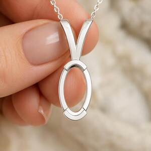 May include: A silver-toned pendant necklace featuring a polished, vertical oval-shaped frame suspended from a delicate chain. The pendant design includes a V-shaped bail that connects to the top of the open oval structure, creating a clean, metallic aesthetic.