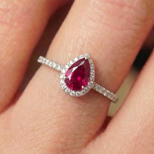 Pode incluir: Um anel com um rubi em forma de pera, apresentando uma pedra preciosa central de cor vermelho profundo rodeada por um halo de pequenos diamantes brancos. A banda é fina e metálica, também incrustada com minúsculos diamantes brancos.