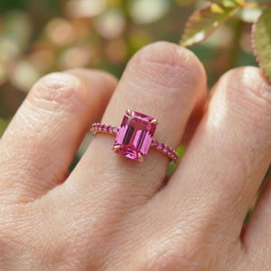 May include: A gold ring featuring a large, rectangular pink gemstone as the centerpiece, flanked by smaller pink stones along the band. The ring is set against a blurred background of green and pink.