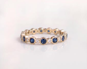 May include: A gold eternity band featuring an alternating pattern of round blue sapphires and clear white diamonds. Each gemstone is set in a circular bezel setting, creating a continuous, uniform design around the entire circumference of the ring.