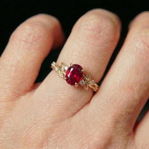 May include: A gold ring featuring a central oval-cut red ruby gemstone. The band is designed with intricate metal leaf motifs on both sides of the stone, accented by small, clear round-cut diamonds embedded within the gold leaf detailing.