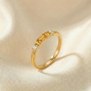 May include: A gold ring featuring a row of five stones set along the top. The center stone is a rectangular yellow baguette, flanked by two round yellow stones and two clear round stones at the outer ends, all set in a polished gold band.