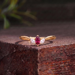 May include: A gold ring featuring a central marquise-cut deep red gemstone flanked by two smaller marquise-cut clear white stones. The band has a curved, V-shaped design that holds the three stones in a horizontal arrangement across the centre.