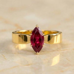 May include: A gold ring featuring a marquise-cut deep red gemstone set in a prong setting. The band is smooth, polished, and wide, providing a solid foundation for the elongated, pointed shape of the central red stone held by six small prongs.