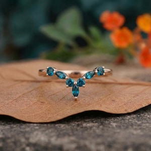 Könnte beinhalten: Ein roségoldener Ring mit geschwungenem Design, besetzt mit tealfarbenen Edelsteinen. Die Edelsteine sind symmetrisch angeordnet, mit einer zentralen Gruppe und flankierenden Steinen. Der Ring wird auf einem braunen Blatt präsentiert.