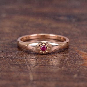 Op de afbeelding: Een roségouden ring met een gladde, gepolijste band en een centrale ronde zetting. De zetting bevat een kleine, ronde, dieprode edelsteen, die wordt omringd door een gegraveerd sterpatroon met acht duidelijke, puntige stralen.