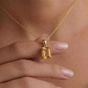 May include: A gold necklace featuring a rectangular emerald-cut yellow citrine gemstone pendant. The pendant is held by a simple gold bail and hangs from a delicate gold chain, showcasing the clear, faceted structure of the stone.