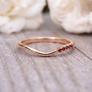 May include: A curved rose gold band ring featuring a row of five small, round, deep red garnet gemstones set along one side of the band. The metal has a polished, smooth finish, and the ring has a distinct wave-like shape to the band's profile.