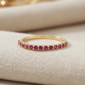 Könnte beinhalten: Ein dünner Goldring mit einer Reihe kleiner, runder, leuchtend pinker Rubine, die entlang der vorderen Hälfte des Bandes gefasst sind. Das Metall hat eine polierte Oberfläche und die Edelsteine sind gleichmäßig in einer Krappenfassung.