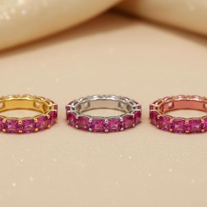 May include: Three eternity bands featuring square-cut vibrant pink stones set in a continuous row. The left ring is yellow gold, the center ring is silver-toned metal, and the right ring is rose gold. Each band displays a uniform, circular design.