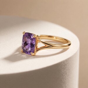 May include: A gold ring featuring a large, cushion-cut purple gemstone set in a four-prong mounting. The band has a split-shank design that tapers as it connects to the central stone, creating a polished and refined appearance on the finger.