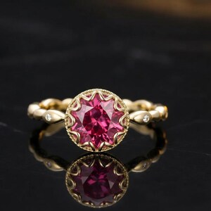 May include: A gold ring with a large, round, pink gemstone at the center. The gemstone is surrounded by a decorative gold setting. The band features a repeating pattern with small, clear stones. The ring is displayed on a reflective black surface.