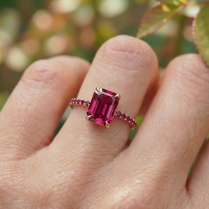 May include: A gold ring featuring a large, rectangular, deep red gemstone as the centerpiece. The band is adorned with smaller, round red gemstones. The ring is set against a blurred background of green and brown.