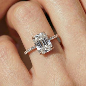 May include: A rose gold ring featuring a large emerald-cut center stone held by four prongs. The band is thin and adorned with a row of small, round, brilliant-cut pavé diamonds extending along the sides of the shank, creating a sparkling effect.