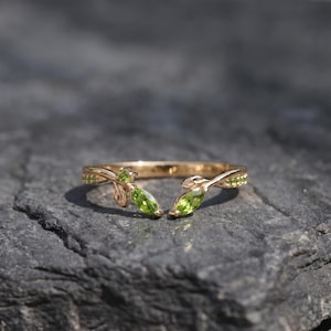 Könnte beinhalten: Ein offener Goldring mit zwei marquise-geschliffenen grünen Peridot-Edelsteinen an den Enden. Die Ringschiene ist mit einem zarten Blattmotiv gestaltet, wobei kleine runde grüne Steine entlang der Metallzweige eingelassen sind.