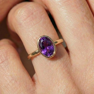 May include: A gold ring with an oval amethyst gemstone. The purple gemstone is set in a bezel setting, and the band is plain gold. The ring is displayed on a finger. The amethyst is a deep purple colour.