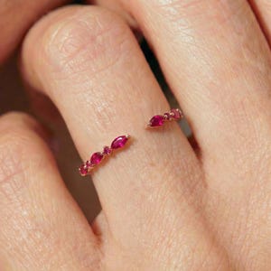 May include: An open-style ring featuring a thin gold band adorned with small, vibrant pink marquise-cut stones. The design includes a series of these stones arranged along the two ends of the open band, creating a delicate and symmetrical appearance.