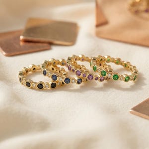 May include: A collection of four gold eternity rings, each featuring a series of bezel-set round gemstones. The rings display alternating stones in black, blue, purple, and green, interspersed with small clear white stones along the gold bands.