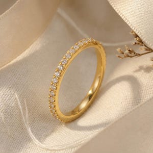 Puede incluir: Un anillo de banda fina de oro con una hilera continua de pequeñas piedras claras de talla redonda engastadas a lo largo de la superficie exterior. El metal tiene un acabado pulido y reflectante, con las piedras dispuestas en una línea.