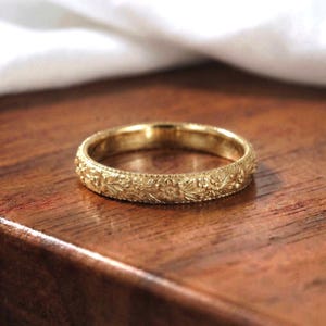 May include: A gold band ring featuring an intricate, continuous floral engraving that wraps around the entire exterior surface. The metal has a polished finish, and the edges of the band are detailed with a fine, beaded milgrain texture.