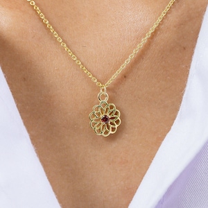 Puede incluir: Un collar de cadena de oro con un delicado colgante que presenta una piedra preciosa roja en el centro de un diseño circular calado.
