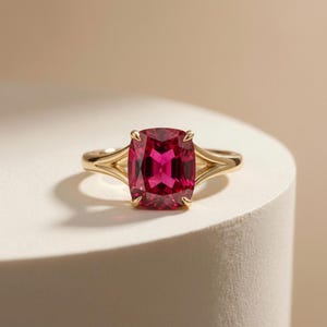 May include: A gold ring featuring a large, rectangular, deep pink gemstone. The ring's band splits into a delicate design on either side of the gemstone, creating an elegant and eye-catching piece of jewelry. The ring is set on a white surface.
