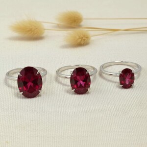 May include: Three silver rings, each featuring a single oval-cut deep red gemstone held by four prongs. The rings are arranged in a horizontal row, showcasing varying sizes of the red stones set on thin, polished silver bands against a neutral background.