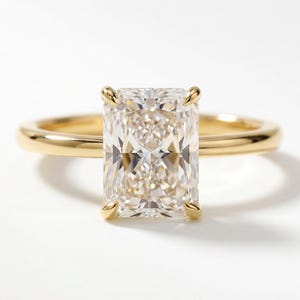 May include: A radiant-cut diamond ring with a gold band. The large, rectangular diamond is set with four prongs, and the gold band has a smooth, polished finish. The ring is displayed on a white surface, highlighting its brilliance.