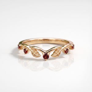 May include: A rose gold ring featuring a V-shaped band adorned with small, carved leaf motifs and five round, deep red garnet gemstones set at intervals along the top curve of the metal band, creating a delicate and symmetrical botanical design.