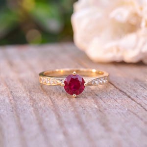 May include: A gold ring featuring a round, deep red ruby set in the center, held by four prongs. The band is crafted from polished yellow gold and is accented with small, clear, round-cut stones embedded along both sides of the shank.