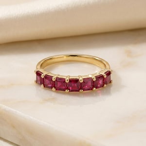 May include: A gold band ring featuring a row of seven square-cut, deep red gemstones set in a prong setting. The ring has a polished metallic finish and the stones are arranged horizontally across the front half of the circular band.