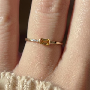 Könnte beinhalten: Ein zarter Goldring mit einem rechteckigen, bernsteinfarbenen Edelstein. Der Ring hat ein dünnes Band mit kleinen, klaren Akzentsteinen auf einer Seite des Edelsteins. Der Ring wird an einem Finger gezeigt.