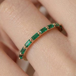 May include: A gold ring with a row of emerald-cut green gemstones. The ring has a thin gold band with small gold beads between each emerald. The ring is shown on a finger.