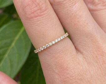 May include: A thin gold band ring featuring a single row of small, round, clear faceted stones set along the top half of the band. The metal has a warm yellow tone, and the stones are arranged in a continuous line, creating a delicate, sparkling appearance.