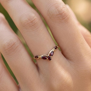 Puede incluir: Un anillo dorado con un diseño curvo, engastado con varias piedras preciosas ovaladas de color rojo intenso. El anillo se lleva en un dedo, mostrando la elegante y delicada artesanía de la joya. Las piedras preciosas miden aproximadamente 0,6 cm de largo.