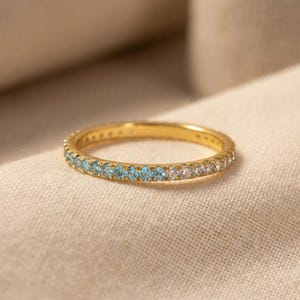 May include: A gold band ring featuring a row of small, round-cut gemstones. The stones are arranged in a gradient pattern, transitioning from light blue on one side to clear, colorless stones on the other side of the circular metal band.
